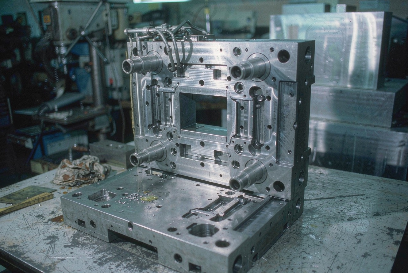 An open steel injection mould on a scratched workshop bench, with hand tools and an oily rag nearby.