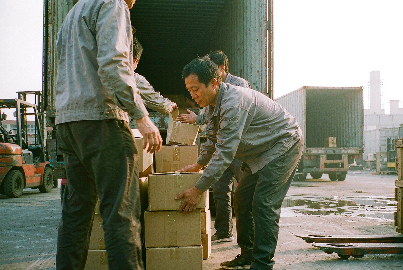 Workers loading sealed cartons into an open shipping container at a factory loading bay