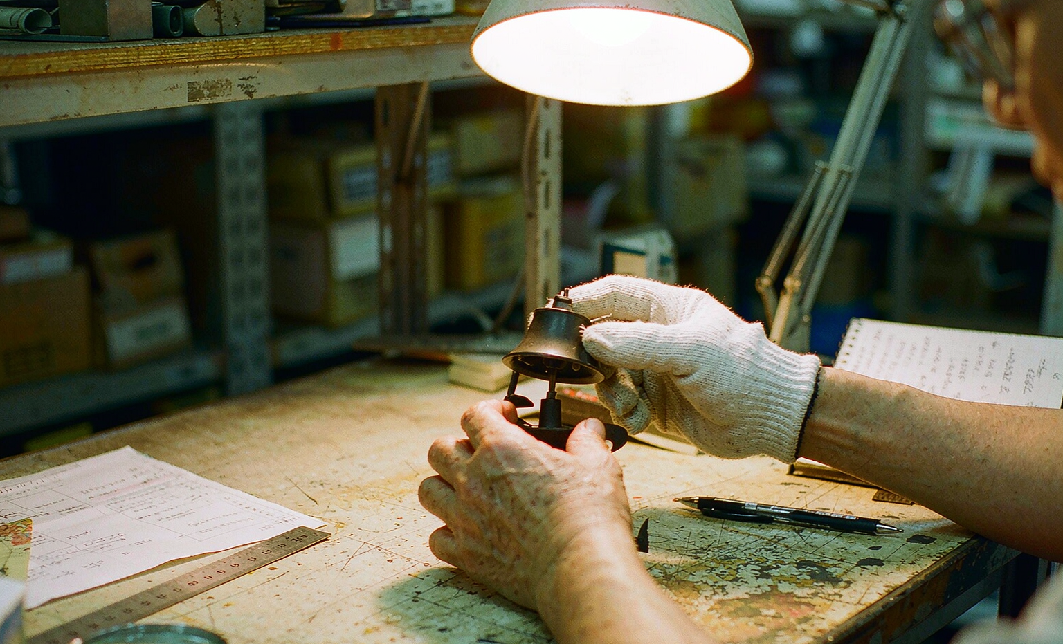 A worker's hands checking a finished product against a ruler and printed spec sheet under a desk lamp