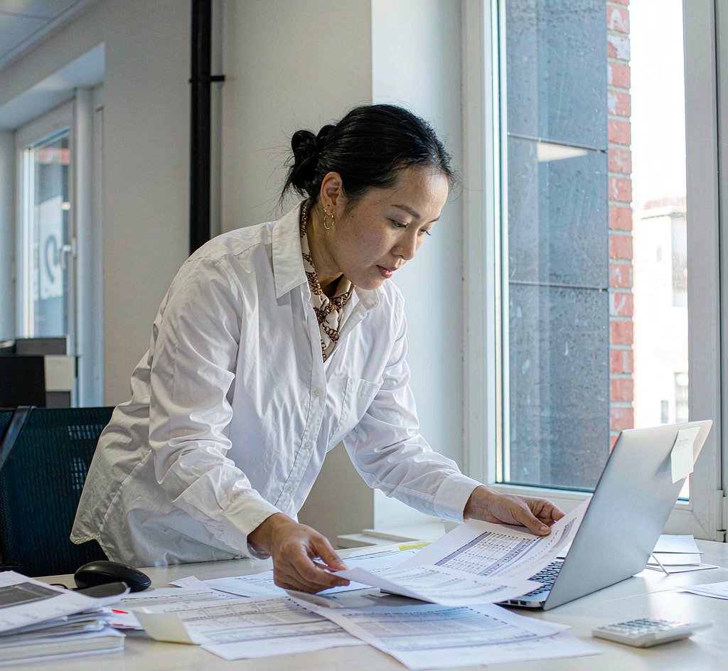 Procurement buyer comparing printed supplier quotations against a spreadsheet on a laptop